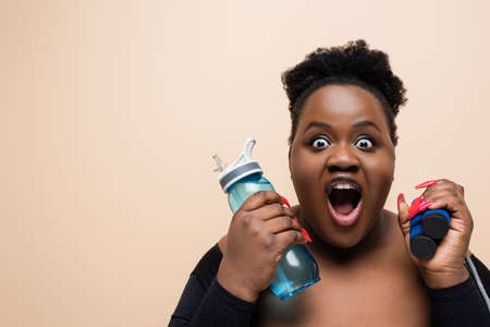 shocked african american oversized woman holding sports bottle and skipping rope isolated on beigeの写真素材