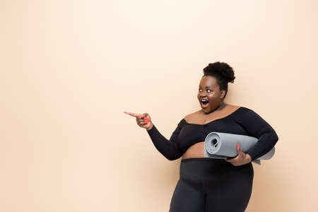 amazed african american overweigh woman in sportswear holding fitness mat and pointing with finger isolated on beigeの写真素材