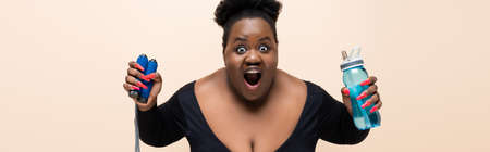 shocked african american plus size woman holding sports bottle and skipping rope isolated on beige, bannerの写真素材