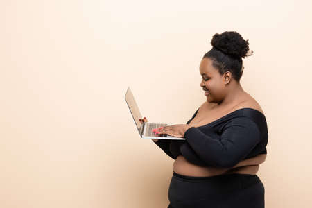 side view of happy african american plus size woman holding laptop isolated on beigeの写真素材