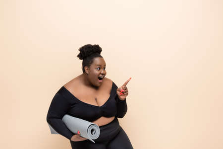 amazed african american plus size woman in sportswear holding fitness mat and pointing with finger isolated on beigeの写真素材