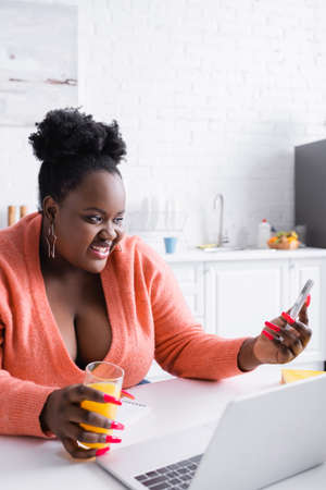 happy african american plus size woman holding glass of orange juice and looking at smartphoneの写真素材