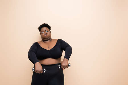 strong african american plus size woman in sportswear holding dumbbells isolated on beigeの写真素材