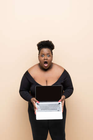shocked african american plus size woman holding laptop isolated on beigeの写真素材