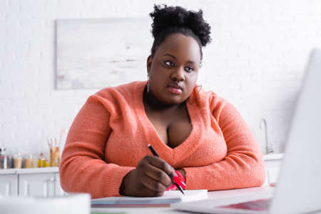 curly african american plus size woman using laptop and holding pen near notebookの写真素材