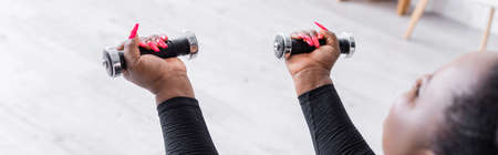 blurred african american plus size woman in sportswear exercising with dumbbells in living room, bannerの写真素材