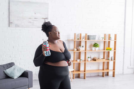 joyful african american overweight woman in sportswear holding sports bottle at homeの写真素材