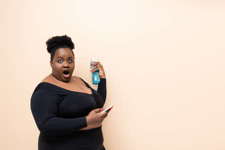 shocked african american plus size woman in sportswear holding sports bottle and smartphone isolated on beigeの写真素材