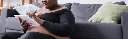 cropped view of pleased african american plus size woman in sportswear using digital tablet in living room, bannerの写真素材