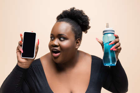 amazed african american plus size woman holding sports bottle and smartphone with blank screen isolated on beigeの写真素材