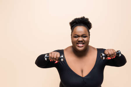happy african american plus size woman in sportswear holding dumbbells isolated on beigeの写真素材