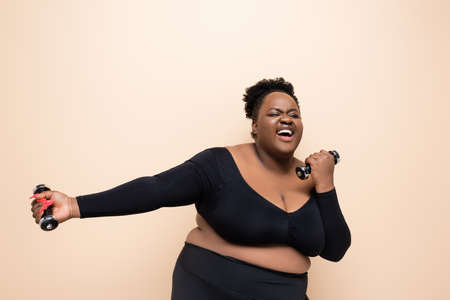 cheerful african american plus size woman in sportswear holding dumbbells isolated on beigeの写真素材