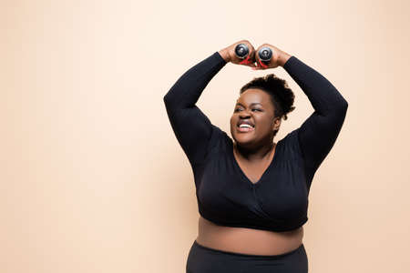 happy african american plus size woman in sportswear holding dumbbells above head isolated on beigeの写真素材