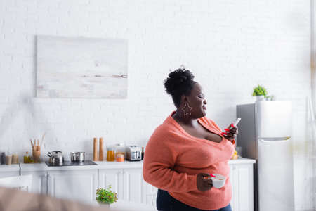 happy african american plus size woman holding on smartphone and cup of coffeeの写真素材