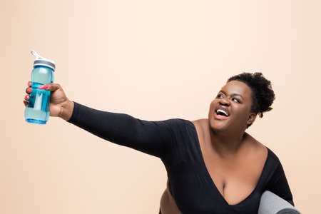 cheerful african american plus size woman in sportswear holding sports bottle isolated on beigeの写真素材
