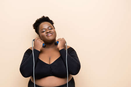 pleased african american plus size woman in sportswear holding skipping rope isolated on beigeの写真素材