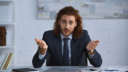 curly businessman in suit pointing with hands and looking at camera in officeの写真素材