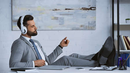 young manager listening music in headphones while sitting with legs on desk in officeの写真素材