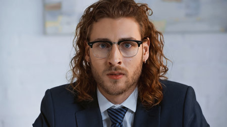 young, curly businessman in eyeglasses looking at camera in officeの写真素材