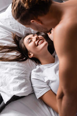 Cheerful woman lying on bed near shirtless boyfriend on blurred foregroundの写真素材