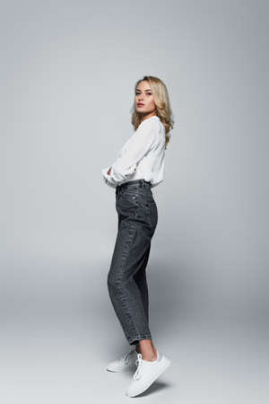 full length view of young, stylish woman looking at camera on grayの写真素材