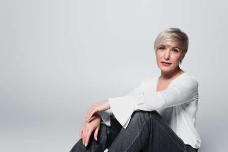 mature woman in white blouse and jeans looking at camera while sitting isolated on gray