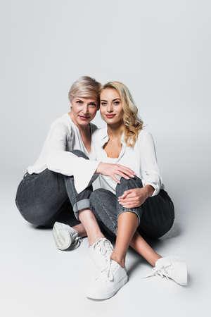 full length view of mother and daughter in white blouses and jeans sitting on grayの写真素材