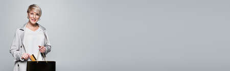 happy woman in stylish outfit putting credit card into shopping bag isolated on gray, bannerの写真素材