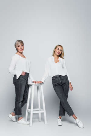 full length view of mature woman with hand in pocket posing near high stool and young daughter on grayの写真素材