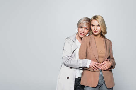 mature, stylish woman hugging adult daughter isolated on gray