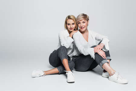 full length view of young woman leaning on shoulder of trendy mother while sitting on gray