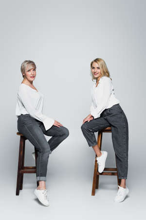 full length view of mother and daughter in white blouses and jeans sitting on high stools on grayの写真素材