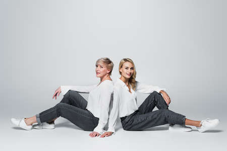 full length view of mother and daughter in trendy clothes sitting back to back on grayの写真素材