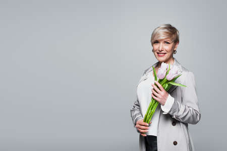 trendy, middle aged woman smiling at camera while holding fresh tulips isolated on grayの写真素材