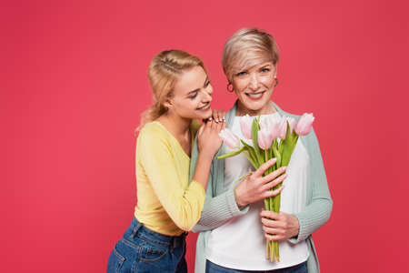 smiling woman leaning on shoulder of joyful mother holding tulips isolated on pink