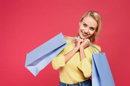 pleased, blonde woman smiling at camera while posing with shopping bags isolated on pinkの写真素材