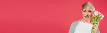 happy, stylish woman looking at camera while holding tulips isolated on pink, banner