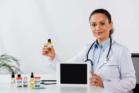 happy african american doctor holding digital tablet with blank screen and cbd bottle near medication on deskの写真素材