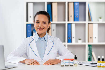 happy african american doctor in white coat smiling near bottles with medical cannabis and cbd lettering on tableの写真素材