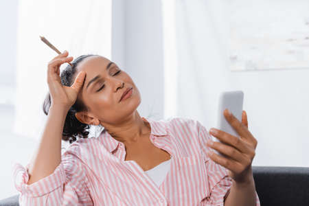 african american woman holding rolled joint with medical cannabis and using smartphone