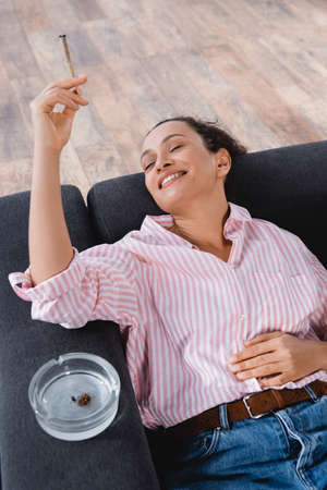 high angle view of pleased african american woman lying on sofa and smoking joint with medical...