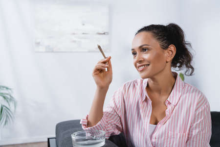 happy african american woman holding rolled joint with medical cannabis near ashtrayの写真素材