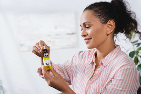 happy african american woman holding bottle with cbd lettering and pipette in living room