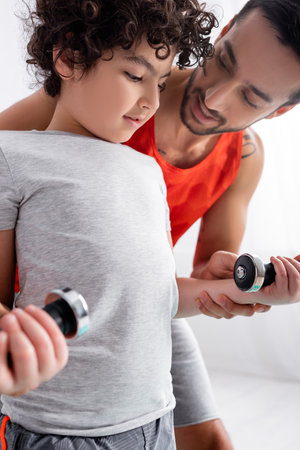 Smiling muslim father holding dumbbells in hands of son at homeの写真素材