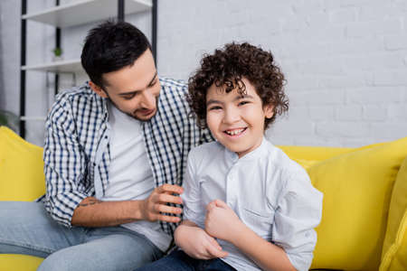 cheerful arabian man tickling joyful son while sitting on sofa at home
