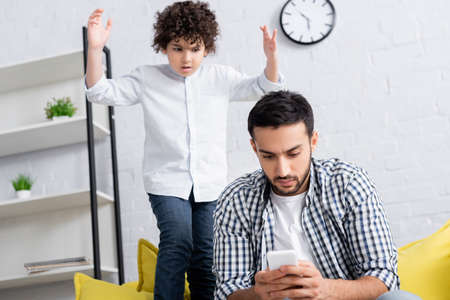 displeased arabian boy standing with raised hands near serious father messaging on smartphoneの写真素材