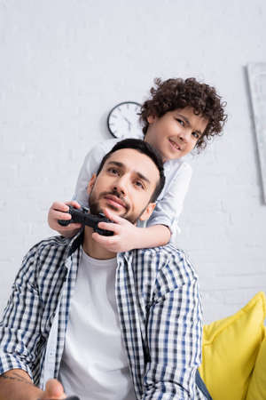 KYIV, UKRAINE - JANUARY 15, 2021: smiling muslim boy playing video game near skeptical fatherの写真素材