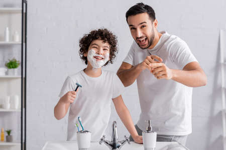 Smiling arabian man pointing with finger near son in shaving foam with razor