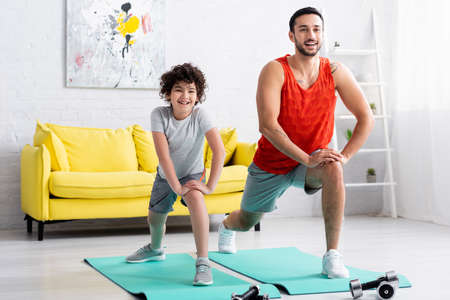 Smiling muslim son and father doing lunges near dumbbells at home
