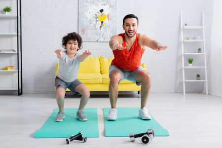 Smiling arabian son and father doing squats on fitness mats near dumbbellsの写真素材